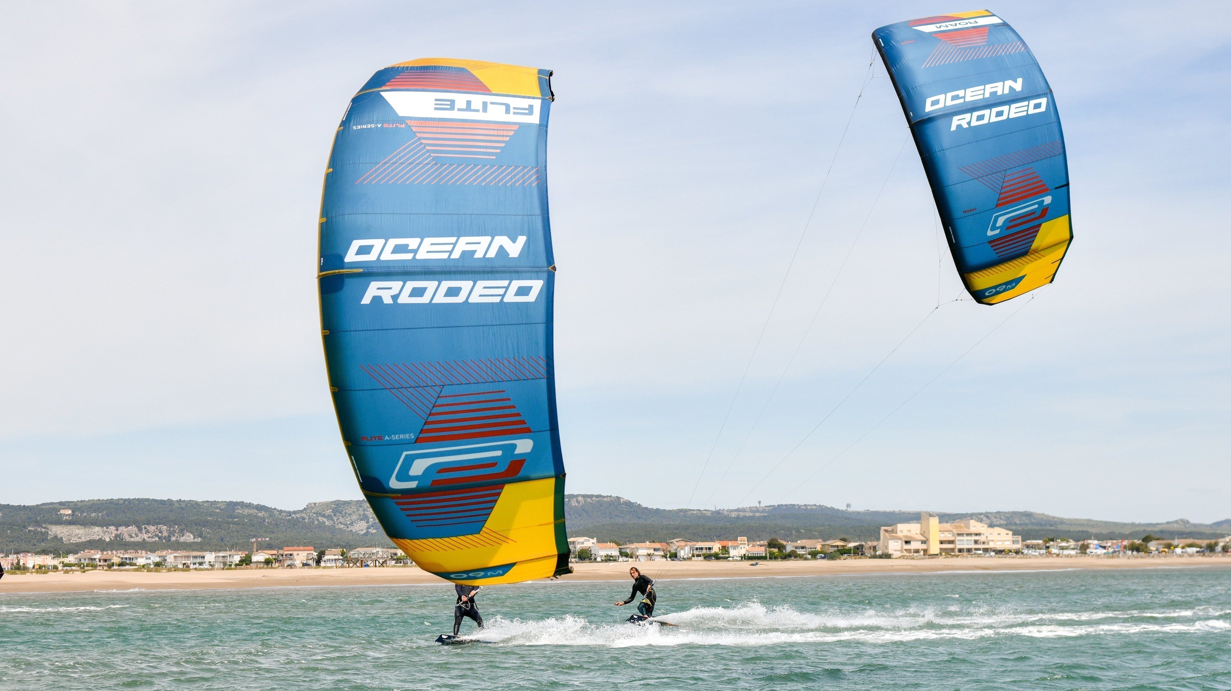 Ocean Rodeo - image shows a wind surfer and a kite surfer on the water looking back towards the beach