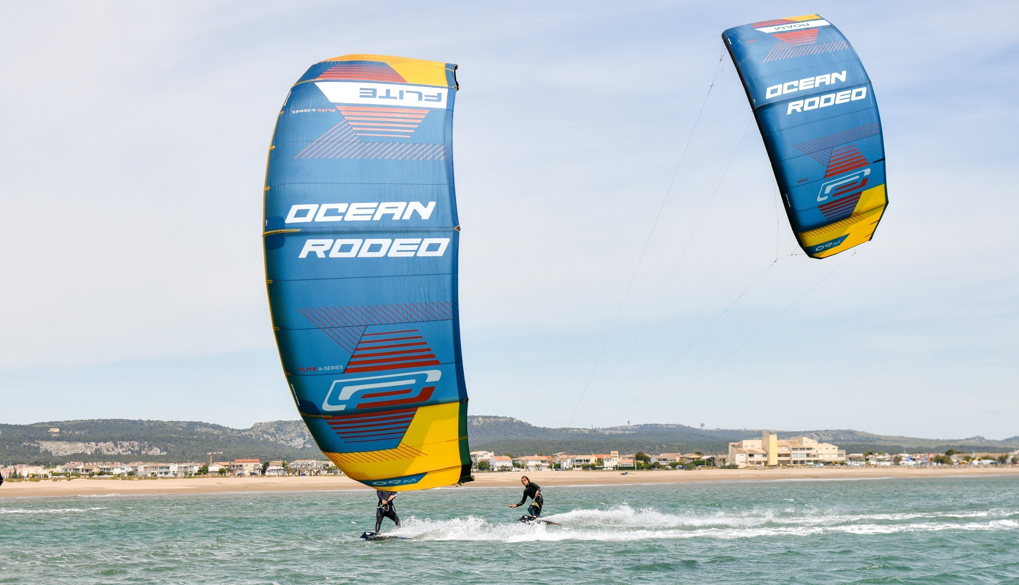 Ocean Rodeo - image shows a wind surfer and a kite surfer on the water looking back towards the beach